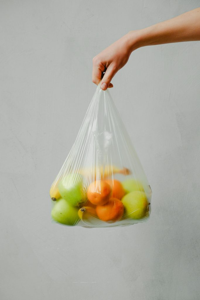 A hand holding a transparent plastic bag full of fresh citrus fruits against a neutral background.